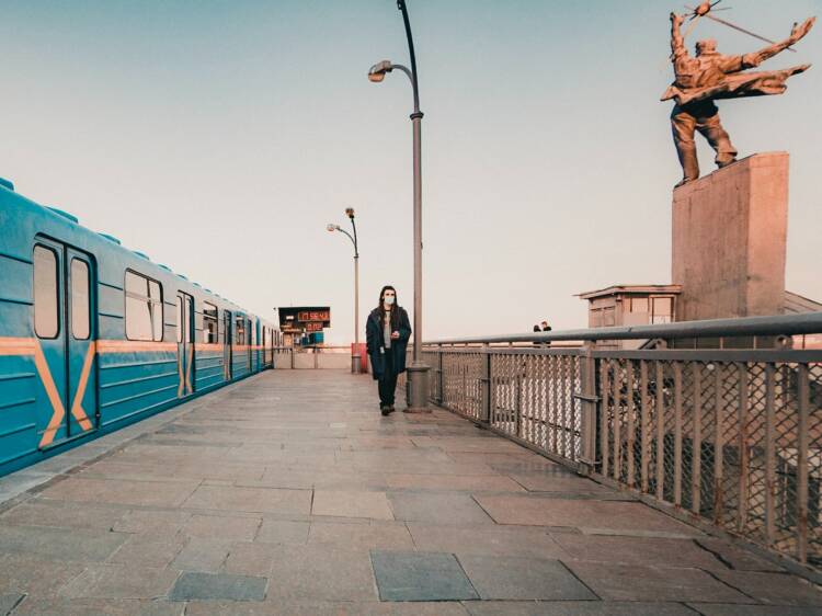Sinikeltainen metrojuna asemalla, laiturilla yksi ihminen. Taustalla aikataulunäyttö ja kuvan oikeassa reunassa ihmishahmoinen patsas.