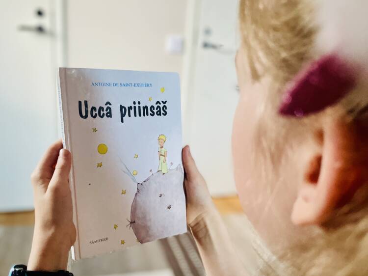 Lapsi pitelee inarinsaamenkielistä Pikku prinssi -kirjaa (Uccâ priinsâš). Kuva on otettu viistosti lapsen takaa.