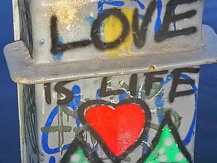 Vanhan sähkökaapin rippeet veden äärellä. Kaapin kylkeen on kirjoitettu ”Love is life”.