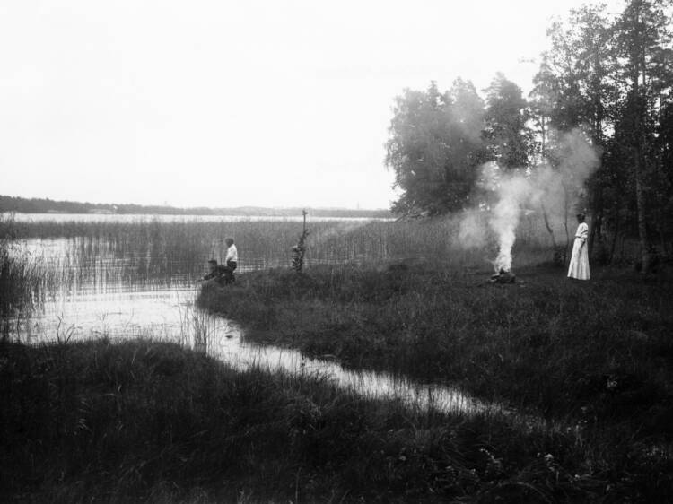 Mustavalkoinen kuva, jossa nuotio veden tuntumassa. Nuotion vieressä nainen, rannassa kaksi lasta.