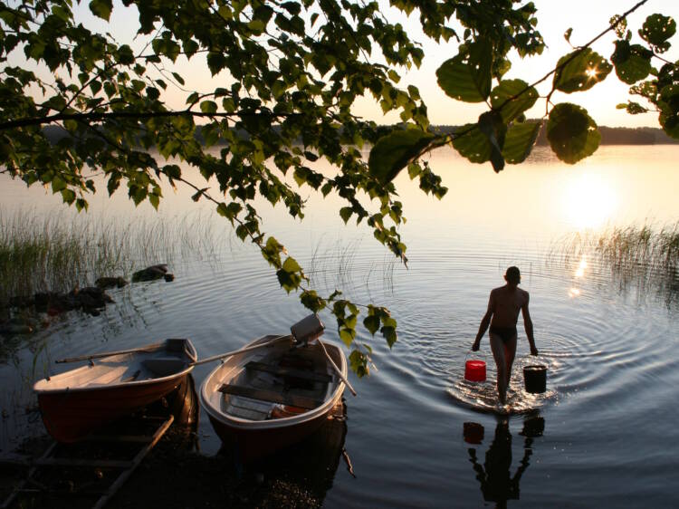 Ihminen kantaa vesistöstä vettä kahdella ämpärillä, vieressä kaksi venettä, taustalla aurinko ja etualalla puiden oksia.