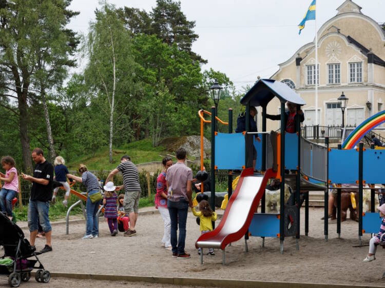 Lapsia ja vanhempia leikkipuistossa kesällä. Taustalla vanha, vaalea kivitalo, puita ja lipputanko, jossa on Ruotsin lippu.