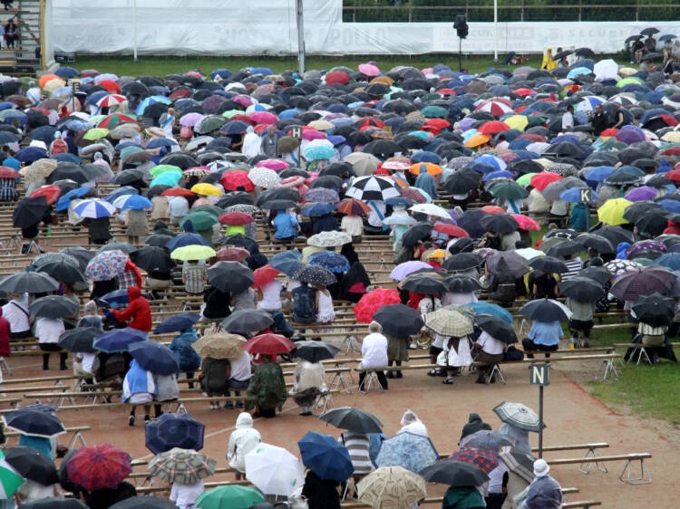 Pitkiä penkkirivejä kuvattuna takaapäin. Penkeillä istuu kymmeniä ihmisiä, joilla on erivärisiä sateenvarjoja.