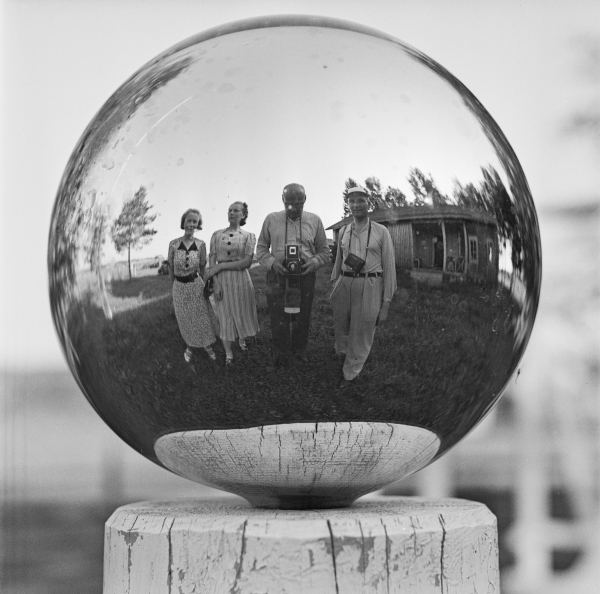 Mustavalkoisessa kuvassa kristallipallo, josta heijastuu neljä ihmistä. Yhdellä heistä kamera, jolla kuva on otettu.