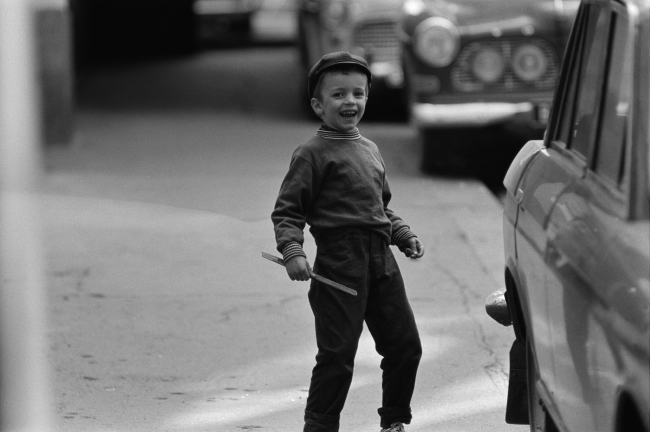 Mustavalkoinen kuva, jos iloinen lapsi kadulla jonkinlainen keppi kädessä. Vieressä pysäköity auto, takana useampi.