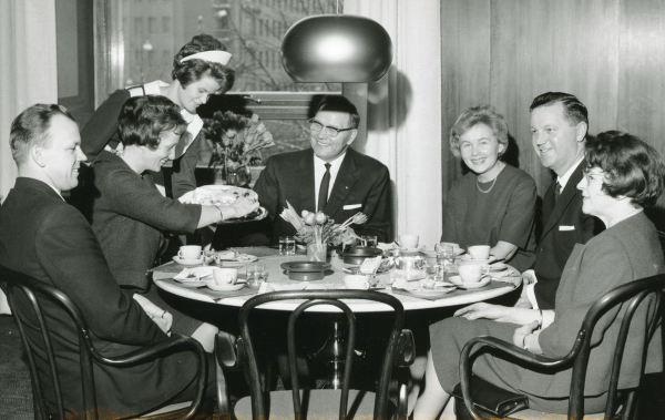 Mustavalkoinen kuva, jossa kuusi ihmistä istuu kahvipöydässä ja tarjoilija ojentaa kakkulautasta.